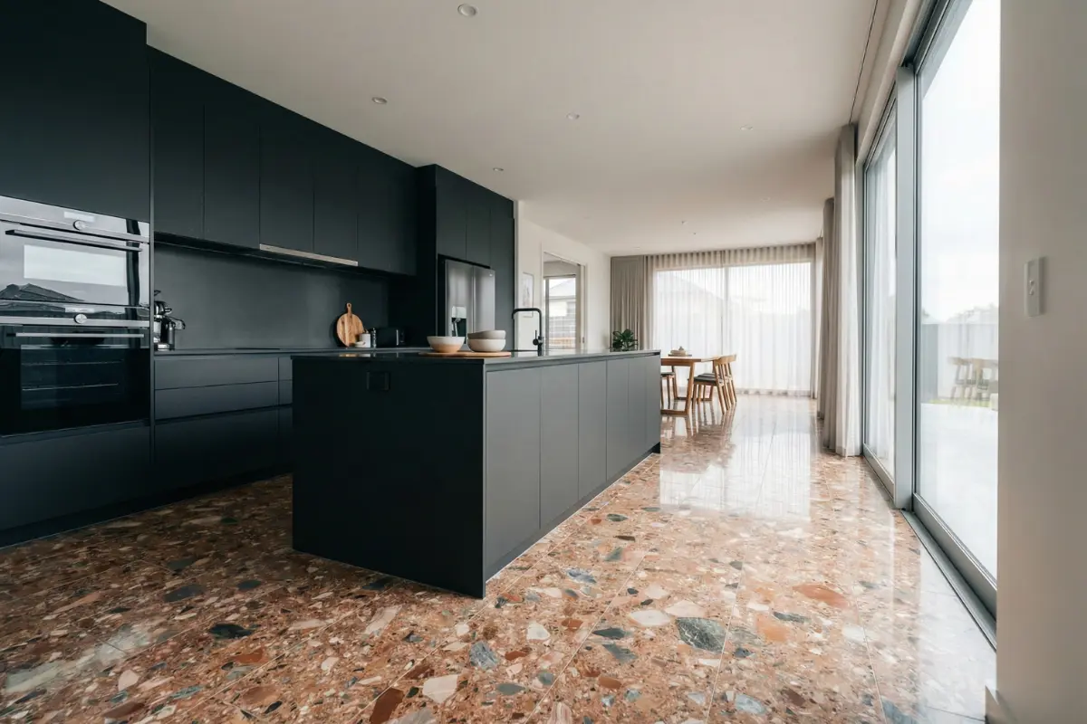 Offene Wohnküche mit glänzenden Terrazzo-Bodenfliesen in Rosé- und Anthrazittönen, schwarzer griffloser Küche mit Kochinsel und Essbereich vor bodentiefen Fenstern – Terrazzo-Fliesen aus Korschenbroich